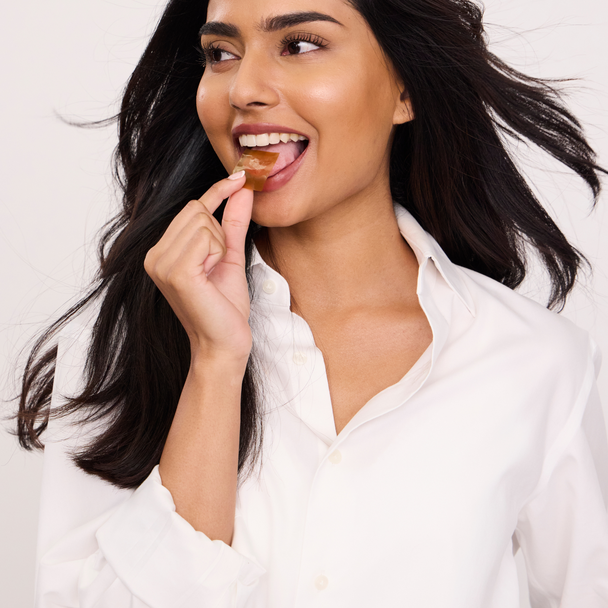 A woman with long dark hair wearing a white button-down shirt is shown in a close-up shot, licking a small brown lip balm or gloss from her finger while looking to the side.